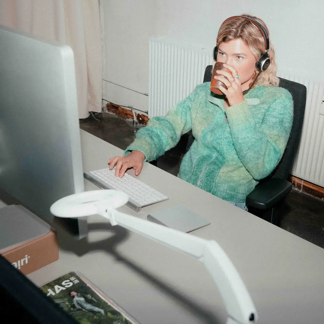 En kvinde i grøn sweater sidder med headset og besvarer kundehenvendelser på computer. Hun drikker en kop kaffe og sidder afslappet og fokuseret.