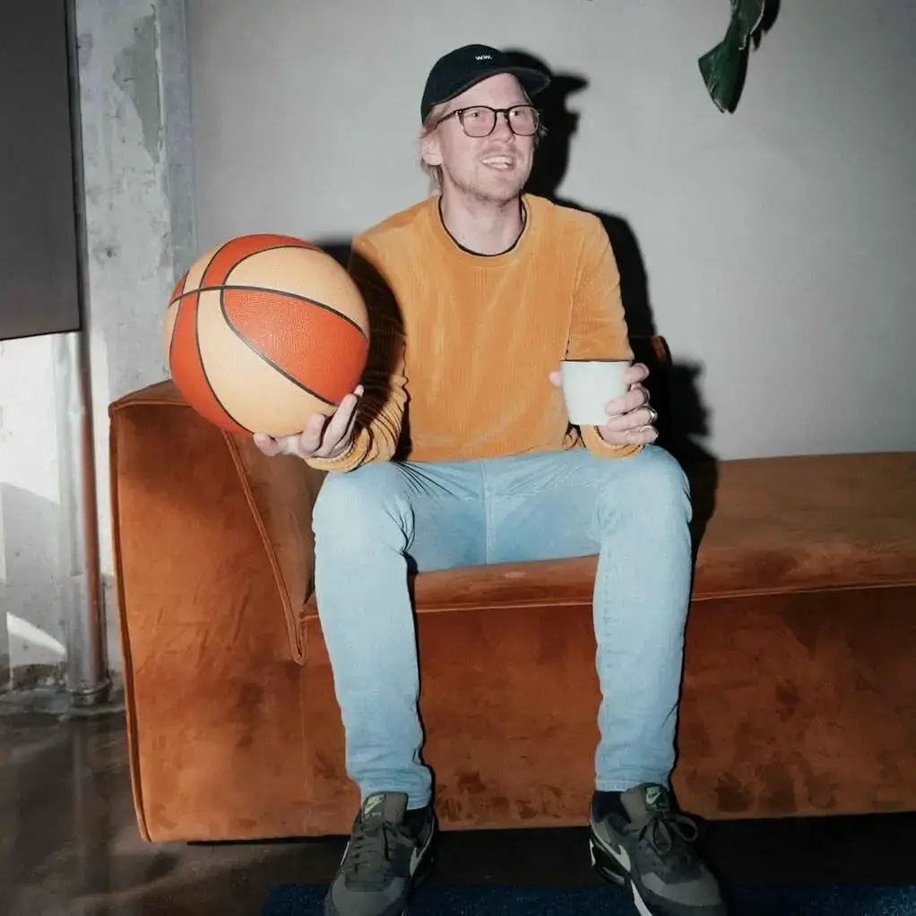 En mand i jeans og sweater sidder på en sofa med en kop kaffe i den ene hånd og en basketball i den anden. han smiler og udstråler overskud.